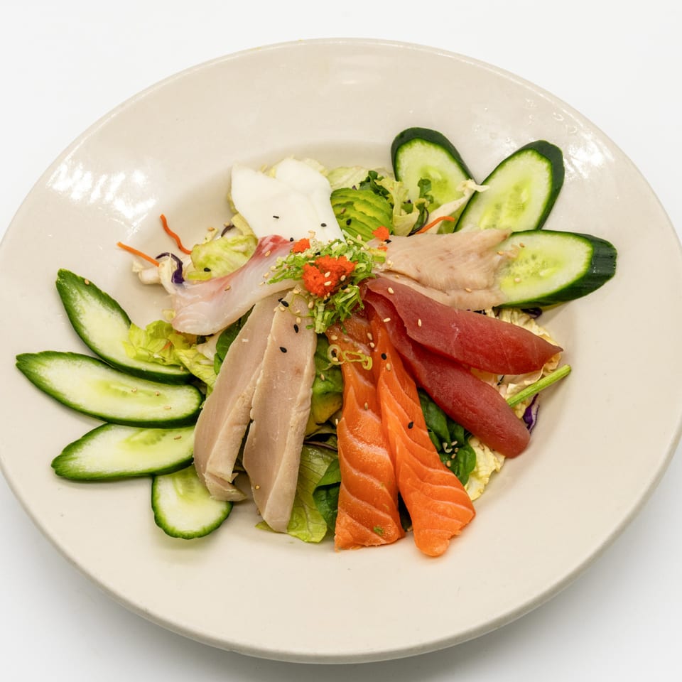 A colorful JooN's Sushi sashimi salad with various fish, cucumbers, and greens on a white plate.