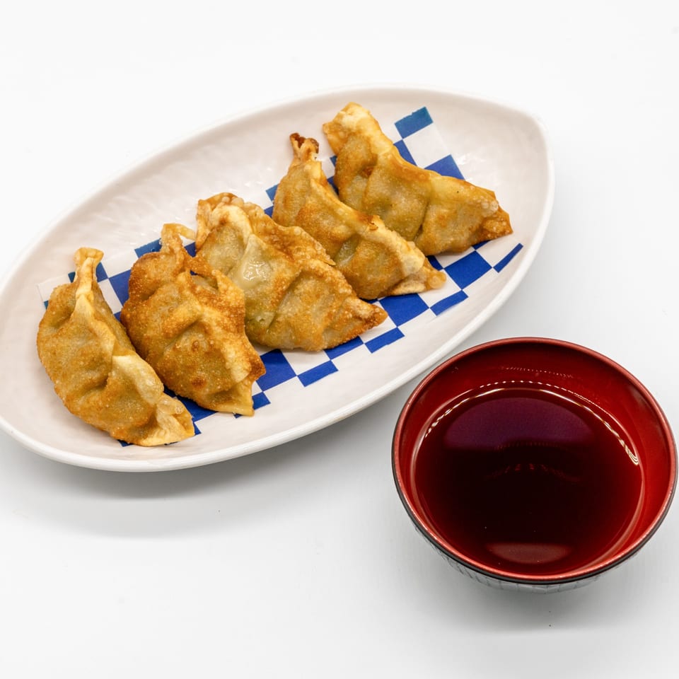 Golden-brown fried gyoza on a white plate with a blue checkered liner, next to a red bowl of sauce.